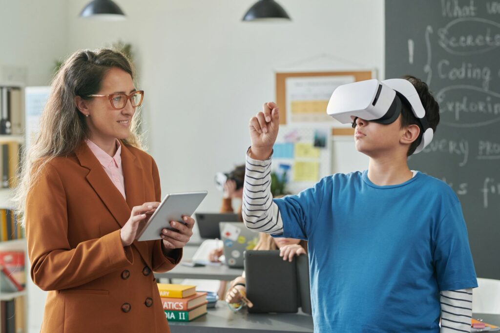 A teacher holding a tablet smiles while watching a student wearing a virtual reality (VR) headset in a classroom. The student, dressed in a blue shirt with striped sleeves, is reaching forward as if interacting with something in the VR environment. In the background, other students are working on laptops and tablets, and a blackboard with handwritten notes is visible.