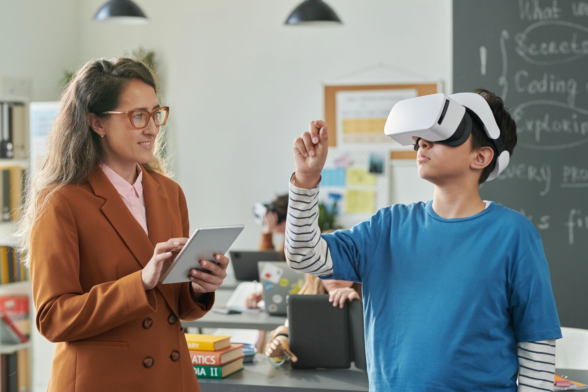 A teacher holding a tablet smiles while watching a student wearing a virtual reality (VR) headset in a classroom. The student, dressed in a blue shirt with striped sleeves, is reaching forward as if interacting with something in the VR environment. In the background, other students are working on laptops and tablets, and a blackboard with handwritten notes is visible.